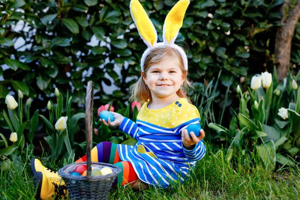 Söt liten småbarn flicka med kaninöron har kul med traditionella påskägg jakt på varm solig dag, utomhus. Glad barn firar familj kristna semester med korg med färgade ägg — Stockfoto