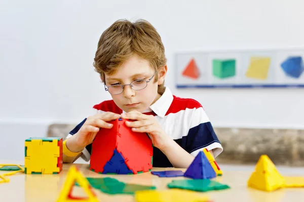小男孩，戴着眼镜，在学校或幼儿园里玩色彩艳丽的塑料元件套件。快乐的孩子建设和创造几何图形，学习数学和几何. — 图库照片