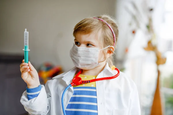 La niña se inyecta. Lindo niño con máscara médica jugando al médico, sosteniendo la jeringa con la vacuna. Concepto de vacunación covid contra el Coronavirus. Juego de rol de niño en casa. —  Fotos de Stock