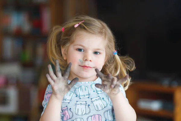 Linda niña adorable aprendiendo pintura con acuarelas. Pequeño dibujo infantil en casa, utilizando pinceles de colores. Niño feliz y saludable con la cara y las manos sucias en casa o en el vivero — Foto de Stock