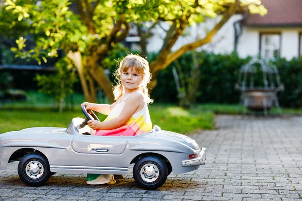 Petite adorable petite fille conduisant une grande voiture de jouet vintage et s'amusant à jouer à l'extérieur. Superbe enfant heureux et en bonne santé profitant d'une chaude journée d'été. Souriant magnifique enfant jouer dans le jardin domestique — Photo