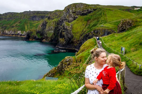 Toddler girl and mother on Carrick-a-Rede Rope Bridge, famous rope bridge near Ballintoy, Northern Ireland on Irish coastline. Family of child and woman on bridge to small island on cloudy day.