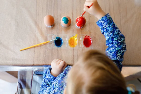 Closeup de menina pequena colorir ovos para a Páscoa. Close-up de criança olhando surpreso com ovos coloridos coloridos, celebrando férias com a família. De cima, rosto não reconhecido. — Fotografia de Stock