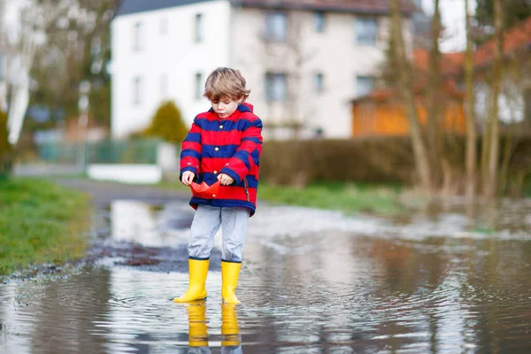 Happy little kid boy in yellow rain boots playing with paper ship boat by huge puddle on spring or autumn day — Stock Photo, Image