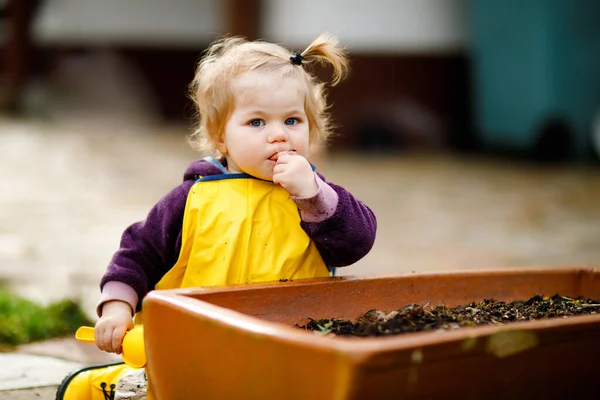Cute adorable toddler girl playing with sand and shovel on spring day. Baby child wearing yellow boots and mud rain puddle pants. Happy girl planting vegetables in spring. — Stock Photo, Image