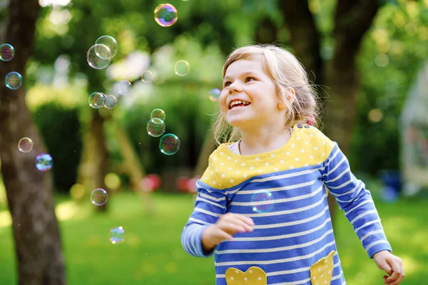 Happy little blonde preschool girl having fun with blowing soap bubble blower. Cute child playing on sunny summer day. Happy active funny healthy kid. Activity for children. — Stock Photo, Image