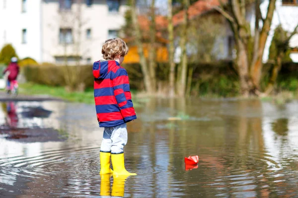 Happy little kid boy in yellow rain boots playing with paper ship boat by huge puddle on spring or autumn day — Stock Photo, Image