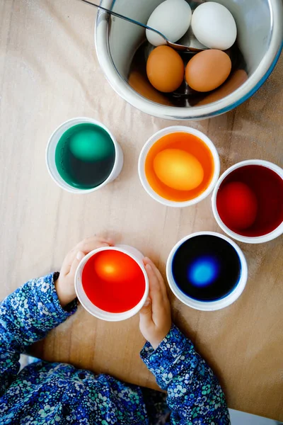 Closeup of little toddler girl coloring eggs for Easter. Close-up of child looking surprised at colorful colored eggs, celebrating holiday with family. From above, unrecognized face. — Stock Photo, Image