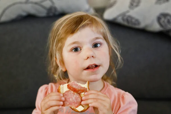 Adorable niñita comiendo sándwich de salami recién preparado en la cocina. El niño feliz come alimentos saludables para el almuerzo o la cena. Aprendizaje infantil. Hogar, guardería, escuela infantil o guardería — Foto de Stock