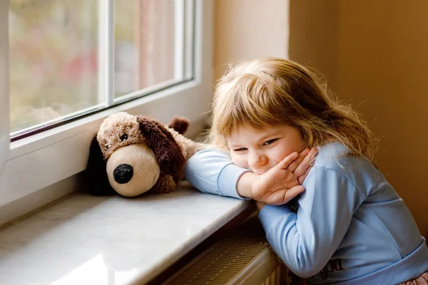 Overstuur peuter meisje zit bij het raam en kijkt uit. Triest huilend kind dat zich ongelukkig voelt. Opsluiting en isolatie concept tijdens de corona virus pandemie tijd. Eenzame kleuter. — Stockfoto