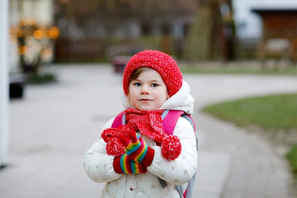 Retrato divertido de una niña preescolar con ropa de invierno. Feliz niño positivo con sombrero rojo y mochila al aire libre en el camino a la escuela infantil, guardería o jardín de infantes. Mañana de invierno. — Foto de Stock