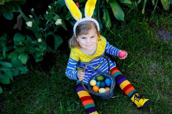 Petite fille mignonne tout-petit avec des oreilles de lapin s'amusant avec des œufs de Pâques traditionnels chassent par une chaude journée ensoleillée, en plein air. Joyeux enfant célébrant des vacances chrétiennes en famille avec panier avec oeuf coloré — Photo