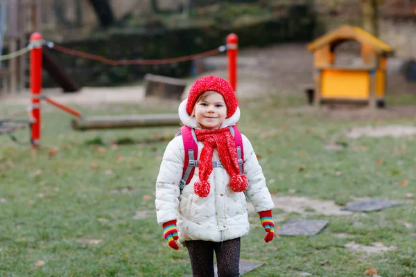 Retrato engraçado de pequena menina pré-escolar em roupas de inverno. Criança positiva feliz com chapéu vermelho e mochila ao ar livre a caminho de playschool, berçário ou jardim de infância. Manhã de inverno. — Fotografia de Stock