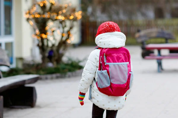 Funny portrait of little preschool girl in winter clothes. Happy positive child with red hat and backpack outdoors on way to playschool, nursery or kindergarten. Winter morning. — Stock Photo, Image