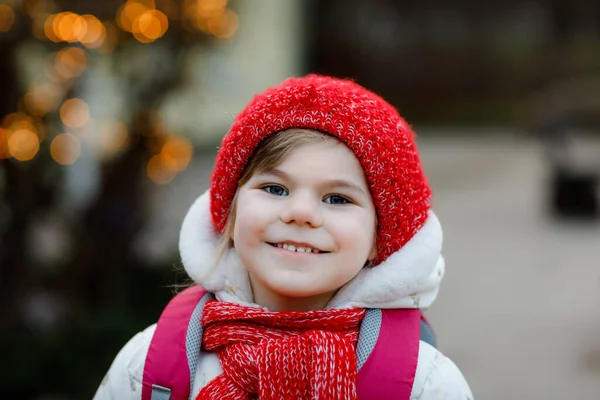 Αστείο πορτραίτο ενός μικρού κοριτσιού προσχολικής ηλικίας με χειμωνιάτικα ρούχα. Ευτυχισμένο θετικό παιδί με κόκκινο καπέλο και σακίδιο σε εξωτερικούς χώρους στο δρόμο για playschool, νηπιαγωγείο ή νηπιαγωγείο. Χειμώνας πρωί. — Φωτογραφία Αρχείου