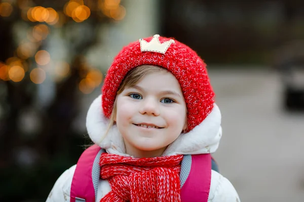 穿着冬衣的学龄前小女孩滑稽的肖像。在去学校、幼儿园或幼儿园的路上，戴着红帽子，背着背包，快乐而积极的孩子。冬季早晨. — 图库照片