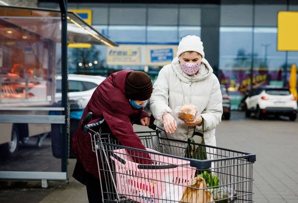 Oudere vrouw en maatschappelijk werker met medisch masker als gevolg van pandemische coronavirus ziekte. Dochter of kleindochter helpen oma met winkelen in de supermarkt, push kar trolley met voedsel, buiten — Stockfoto