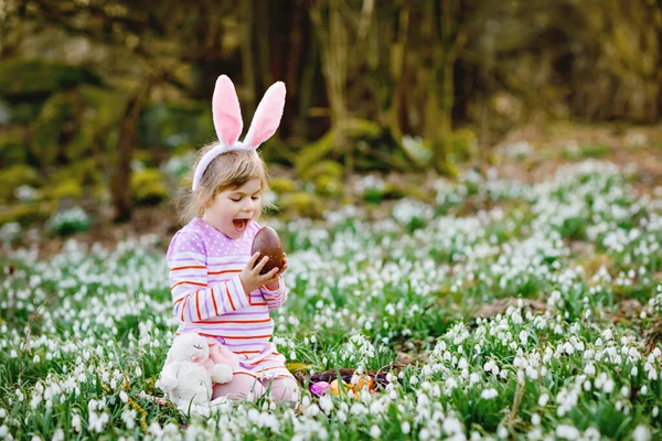 一个小女孩，带着复活节兔子的耳朵，在阳光明媚的春天的森林里打猎。可爱快乐的孩子，有很多雪花，巨大的巧克力蛋和彩蛋. — 图库照片