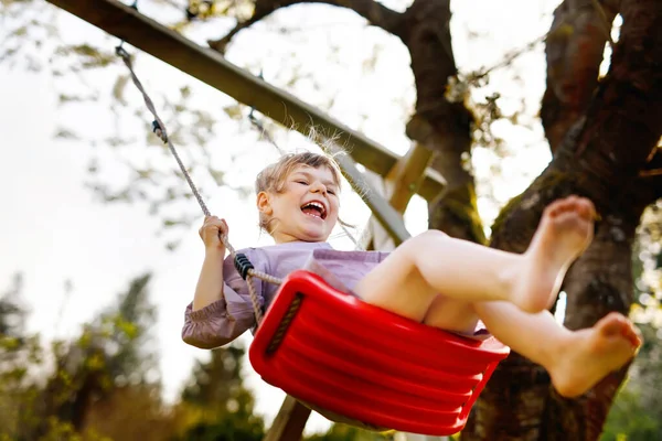 Glad liten småbarn flicka som har kul på swing i hushållsträdgården. Leende positiva friska barn swingon solig dag. Förskoleflickan skrattar och gråter. Aktiv fritid och aktivitet utomhus. — Stockfoto