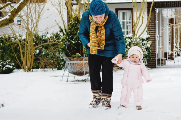 Adorable little baby girl making first steps outdoors in winter through snow. Cute toddler learning walking. Mother holding child on hand. Daughter and mum walk together. Happy family — Stock Photo, Image