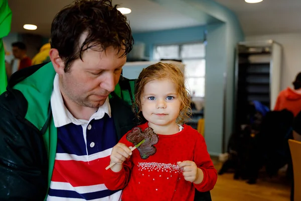 Cute toddler girl and father with cloverleaf lollipop in Irish pub or cafe. Child and dad, young man resting with parents and eating unhealthy sweets. Family making vacation in Ireland. — Stock Photo, Image