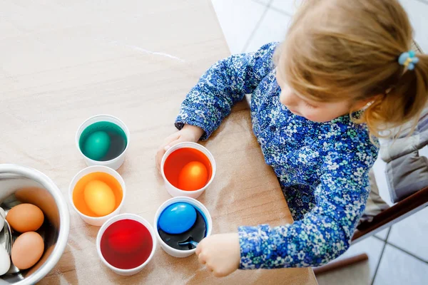 Primer plano de la niña pequeña para colorear huevos para Pascua. Primer plano del niño que mira sorprendido por los huevos de colores de colores, celebrando la fiesta con la familia. Desde arriba, cara no reconocida. — Foto de Stock