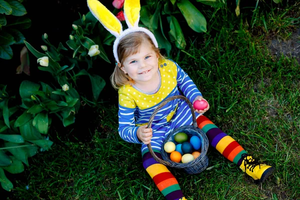 Petite fille mignonne tout-petit avec des oreilles de lapin s'amusant avec des œufs de Pâques traditionnels chassent par une chaude journée ensoleillée, en plein air. Joyeux enfant célébrant des vacances chrétiennes en famille avec panier avec oeuf coloré — Photo