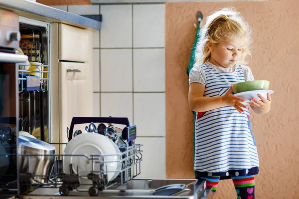 A kis aranyos, aranyos kislány segít kipakolni a mosogatógépet. Vicces, boldog gyerek áll a konyhában, edényeket tart és tálat tesz a fejére. Egészséges gyerek otthon. Gyönyörű segítő szórakozás — Stock Fotó