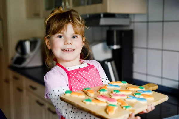 Cute little toddler girl holding fresh baked homemade Easter or spring cookies at home indoors. Adorable blond child with apron with bunny and carrot cookie in domestic kitchen. — Stock Photo, Image