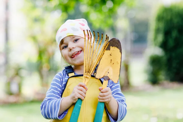 Roztomilé malé batole dívka drží zahradní nářadí v rukou. Roztomilé dítě se učí zahradničení, výsadbě a pěstování zeleniny v domácí zahradě. Kluk s lopatou a hráběmi. Ekologie, organické potraviny. — Stock fotografie
