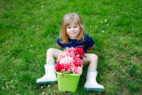 Linda adorable niña pequeña con muchas flores de peonía rosa en cubo. Feliz niño sonriente en el jardín doméstico en el cálido día de primavera o verano. —  Fotos de Stock