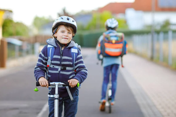 Due scolaretti in casco di sicurezza a cavallo con scooter in città con zaino nella giornata di sole. Bambini felici in vestiti colorati in bicicletta sulla strada per la scuola. — Foto Stock