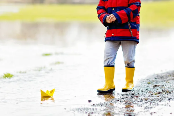 Happy little kid boy in yellow rain boots playing with paper ship boat by huge puddle on spring or autumn day — Stock Photo, Image