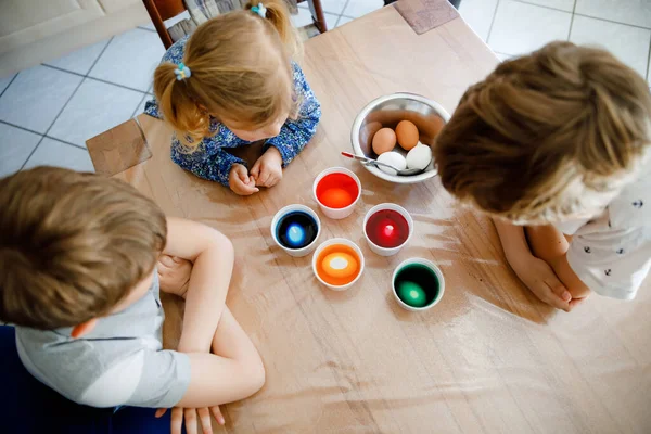 Menina criança animada e dois meninos crianças colorir ovos para a Páscoa. Três crianças, irmãos olhando surpresos com ovos coloridos, celebrando férias com a família. De cima, rostos não reconhecidos — Fotografia de Stock