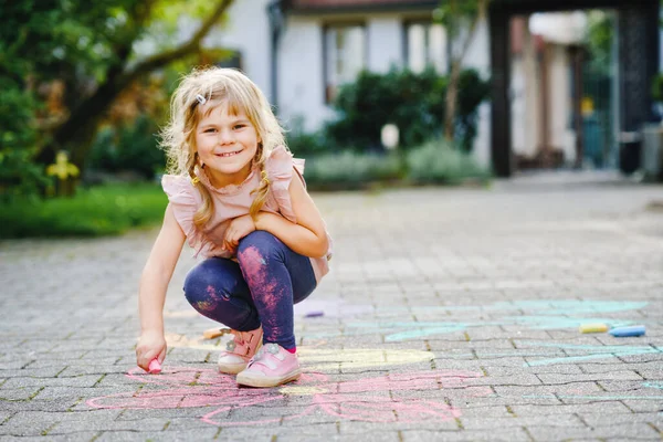 Little preschool girl painting with colorful chalks flowers on ground on backyard. Positive happy toddler child drawing and creating pictures on asphalt. Creative outdoors children activity in summer. — Stock Photo, Image