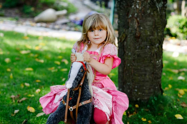 Malá školačka objímající se s houpající se koňskou hračkou. Šťastné dítě v princezně šaty na slunné letní den v zahradě. Dívka v lásce se svým oblíbeným starým veteránem hračka zvíře. — Stock fotografie