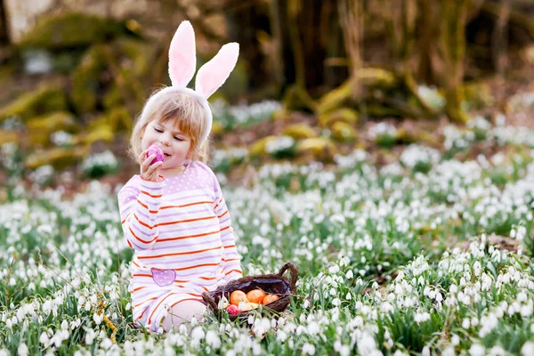 Petite fille aux oreilles de lapin de Pâques faisant la chasse aux œufs dans la forêt de printemps par une journée ensoleillée, en plein air. Mignon enfant heureux avec beaucoup de fleurs de chute de neige et des œufs colorés. Printemps, concept de vacances chrétiennes. — Photo