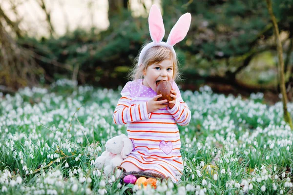 Holčička s velikonočními králičími ušima, která loví vajíčka v jarním lese za slunečného dne venku. Roztomilé šťastné dítě se spoustou zasněžených květin, jíst obrovské čokoládové vejce a barevné vejce. — Stock fotografie
