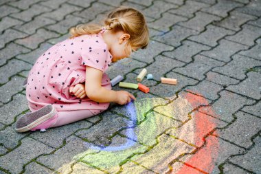 Küçük anaokulu kızı arka bahçede renkli tebeşirlerle gökkuşağını boyuyor. Çocuk resim çizerken ve resim çizerken çok mutlu. Yazın yaratıcı açık hava aktivitesi.