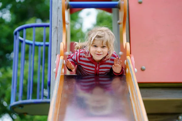 En liten förskoletjej som leker på lekplatsen. Glada småbarn klättra och ha kul med sommaren utomhus aktivitet. Flicka slinka ner bilden. — Stockfoto