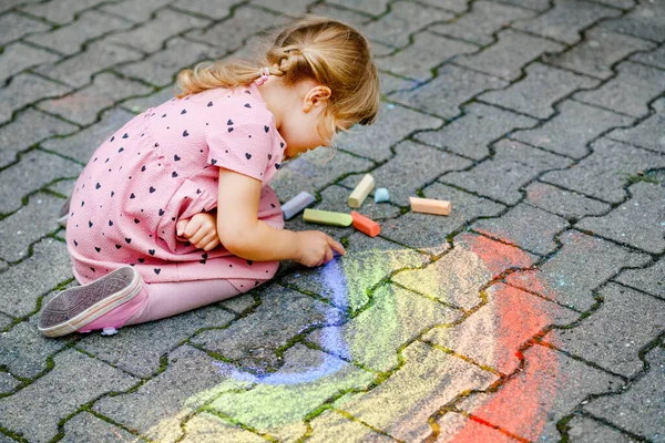 Маленькая дошкольница рисует радугу с разноцветными мелом на земле на заднем дворе. Счастливый малыш рисует и создает картины. Творческая деятельность на открытом воздухе летом. — стоковое фото