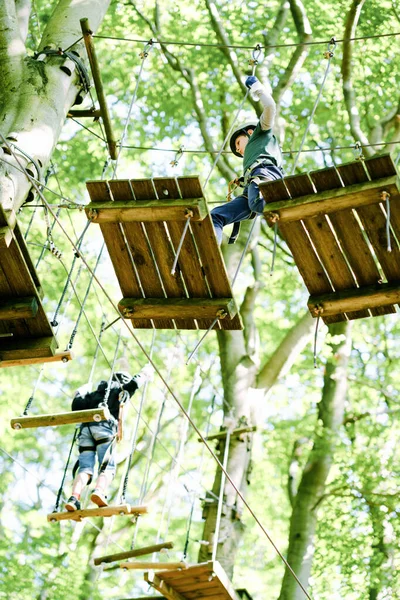 Duas crianças no parque de aventura florestal. Crianças meninos de capacete sobe em trilha corda alta. Habilidades de agilidade e escalada centro de diversões ao ar livre para crianças. Atividades ao ar livre para crianças e famílias. — Fotografia de Stock