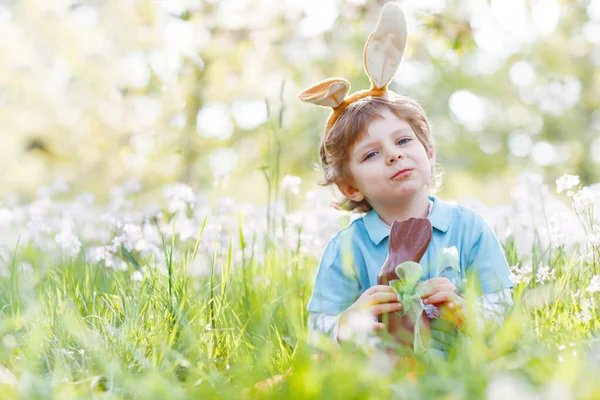 Cute little kid boy with Easter bunny ears celebrating traditional feast. Happy child eating chocolate rabbit fugure — Stock Photo, Image