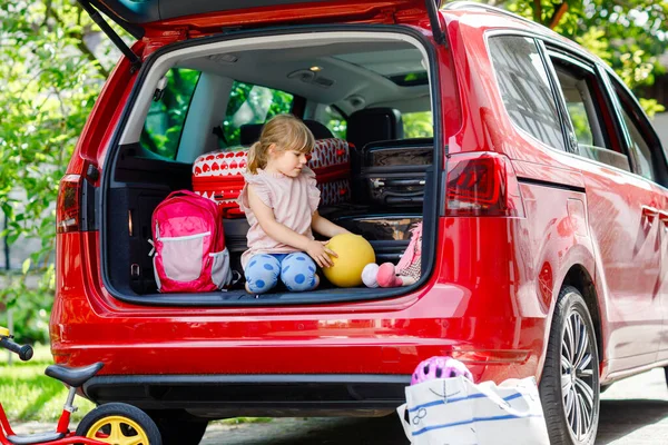 Roztomilá malá školačka sedící v kufru auta před odjezdem na letní dovolenou s rodiči. Šťastné aktivní dítě s kufry, koly a hračkami na rodinné cestě, výlet — Stock fotografie