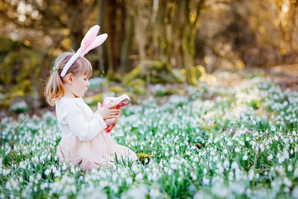 可爱的小女孩，带着复活节兔子的耳朵，在阳光灿烂的春天的森林里，在户外吃着令人窒息的人形食物。带着许多雪花的可爱的快乐的孩子。春天，基督教节日的概念. — 图库照片