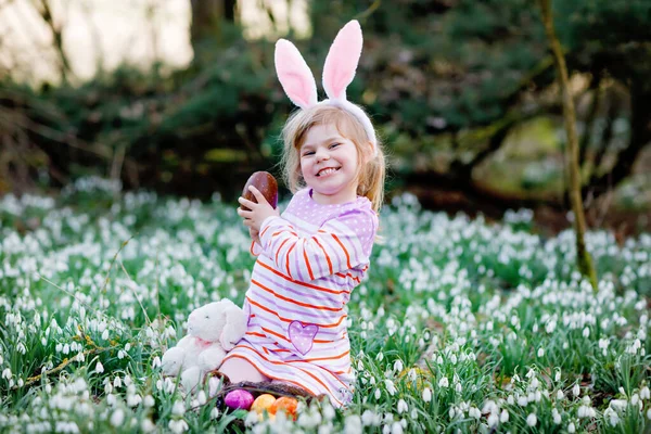 一个小女孩，带着复活节兔子的耳朵，在阳光明媚的春天的森林里打猎。可爱的孩子，有很多雪花，吃着巨大的巧克力蛋和彩蛋. — 图库照片