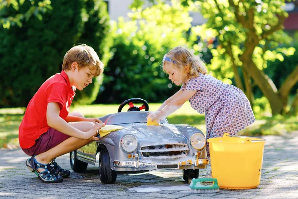 Zwei glückliche Kinder waschen im Sommergarten im Freien ein großes altes Spielzeugauto. Bruder Junge und kleine Schwester Kleinkind Mädchen reinigen Auto mit Seife und Wasser, haben Spaß beim Planschen und Spielen mit Schwamm. — Stockfoto