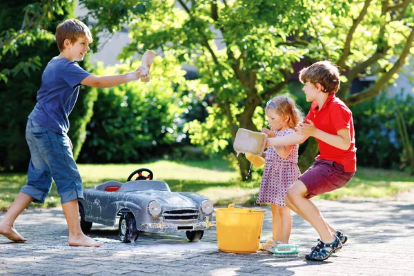 Três crianças felizes lavando grande carro de brinquedo velho no jardim de verão, ao ar livre. Dois meninos e pequena menina limpando carro com água e sabão, se divertindo com espirrar e brincar com esponja. — Fotografia de Stock