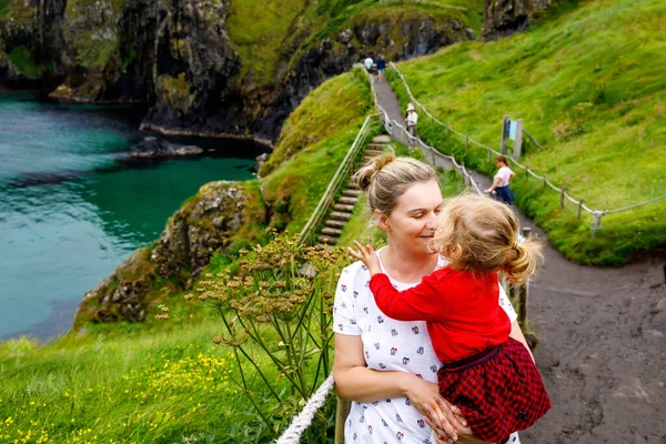Batole dívka a matka na Carrick-a-Rede laně most, slavný lanový most u Ballintoy, Severní Irsko na irském pobřeží. Rodina dítěte a ženy na mostě na malý ostrov v zamračený den. — Stock fotografie