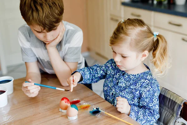Spännande liten småbarn flicka och äldre bror, skolpojke färga ägg för påsk. Två söta barn, syskon tittar förvånad över färgglada ägg, firar semester med familjen. — Stockfoto